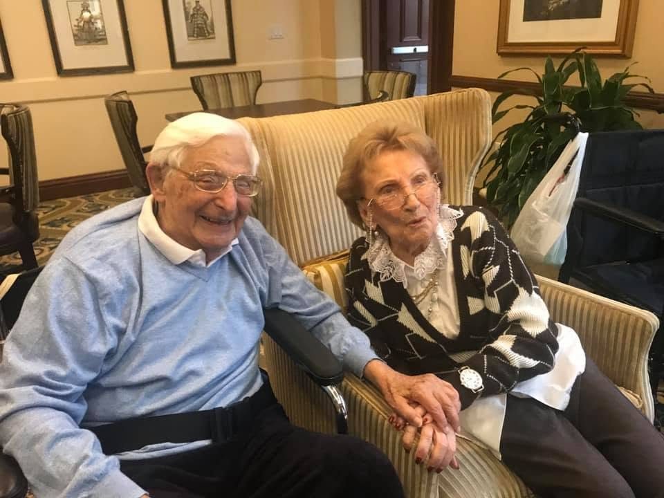 An elderly couple seated together on a sofa, holding hands